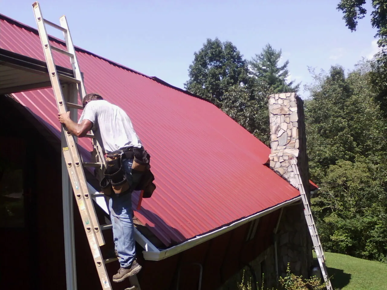 Roofer on a ladder beside a red metal roof and stone chimney — Soffit & Fascia Repair by Roofing Brothers in Charlton
